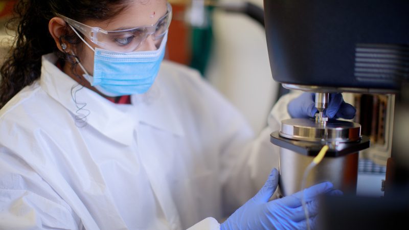 A woman in lab coat and face mask works in a lab.