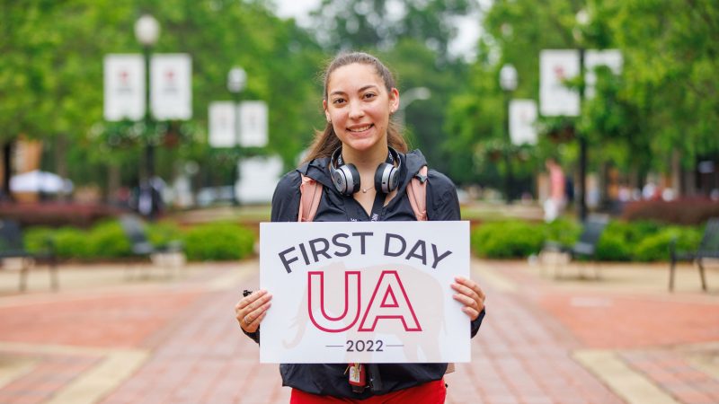 a young woman holds up a sign that says "First Day UA 2022"