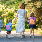 A woman walks with three little girls who are wearing backpacks.