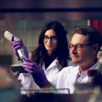 Two UA chemists work in the lab in white coats and gloves.