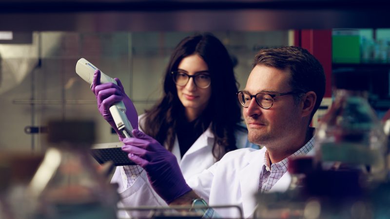 Two UA chemists work in the lab in white coats and gloves.