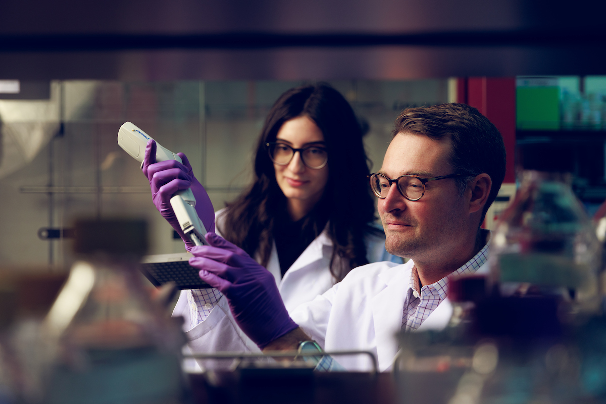 Two UA chemists work in the lab in white coats and gloves.