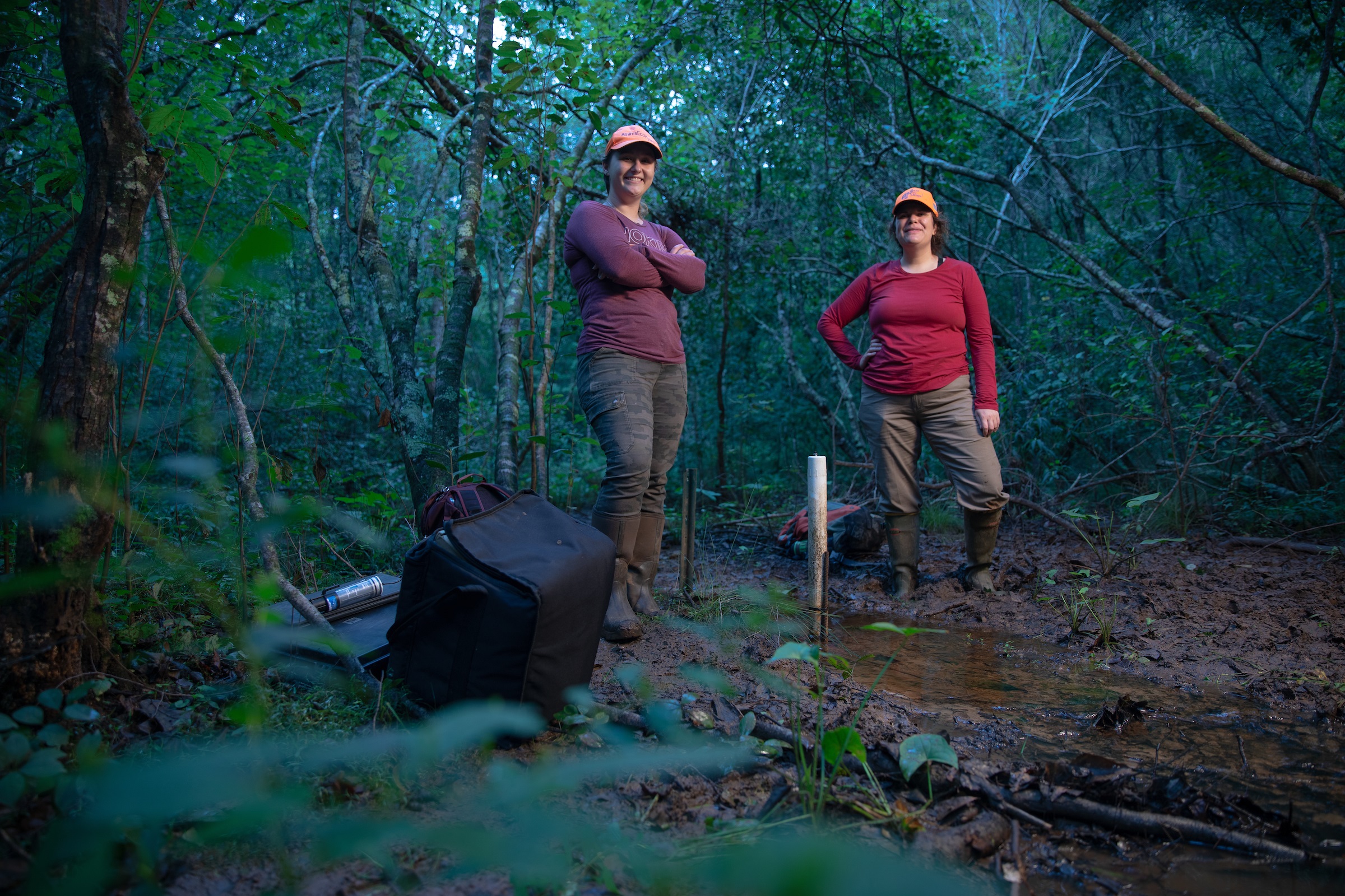Two researchers in mud boots pose for a photo by a forest stream.