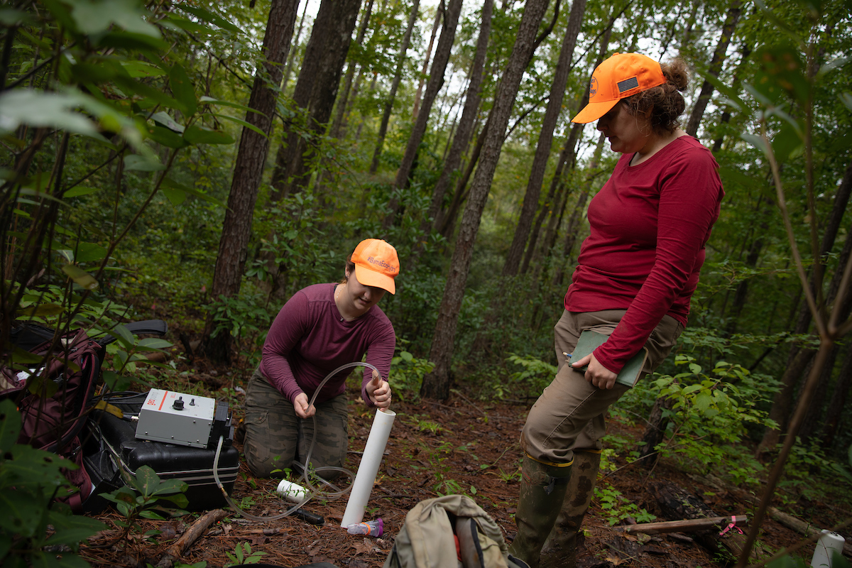 Two researchers surrounded by a equipment in a forest, run a clear hose down a pipe into the ground.