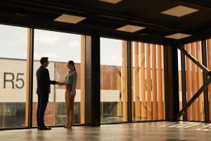 Wide angle view of real estate agent shaking hands with client while standing in empty office building interior lit by sunlight.