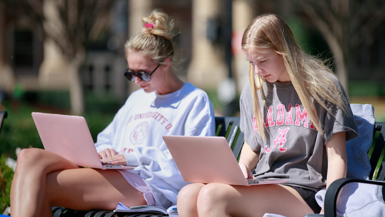 two young women work on their laptops on a bench at the Quad
