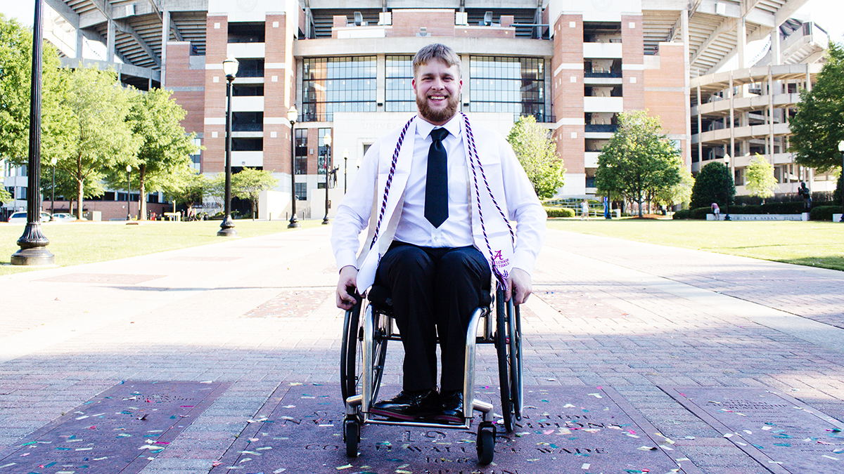 A man wearing a white graduation gown is sitting in a wheelchair in front of Bryant Denny Stadium.