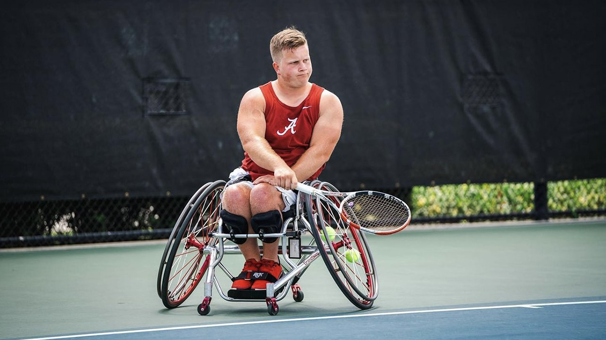 A young blonde man is playing tennis while sitting in a wheelchair.