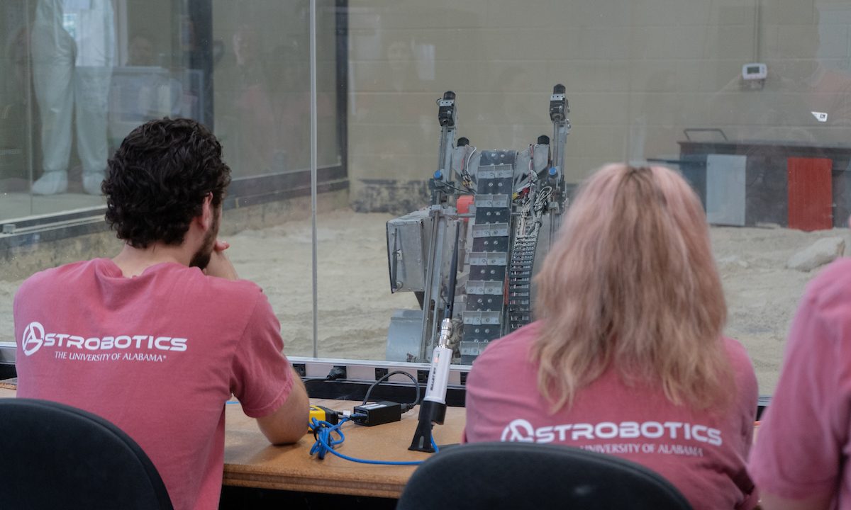 Students at The University of Alabama watch as a rover-sized robot prepares to dig inside a glass room.