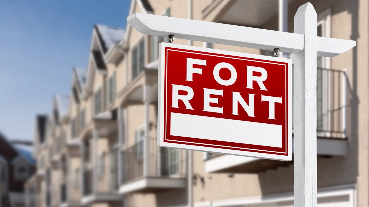 For Rent Real Estate Sign In Front of a Row of Apartment Condominiums Balconies and Garage Doors.