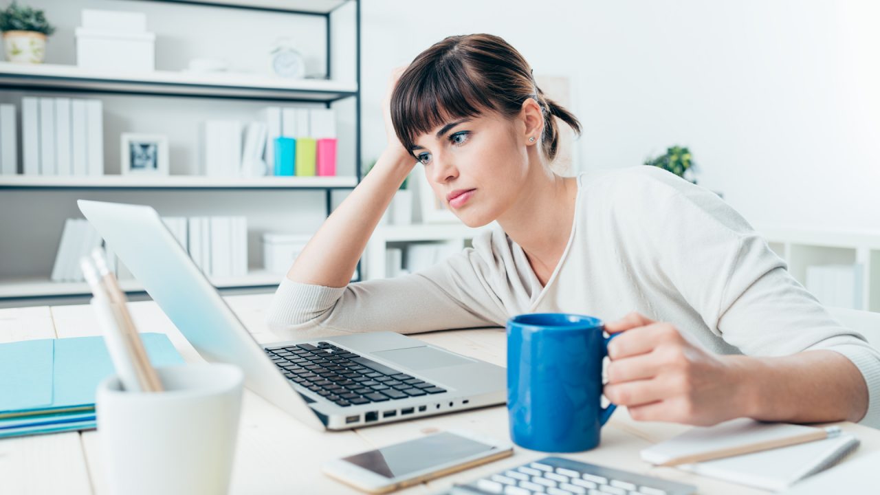 A tired woman sitting in front of a computer holding a coffee cup