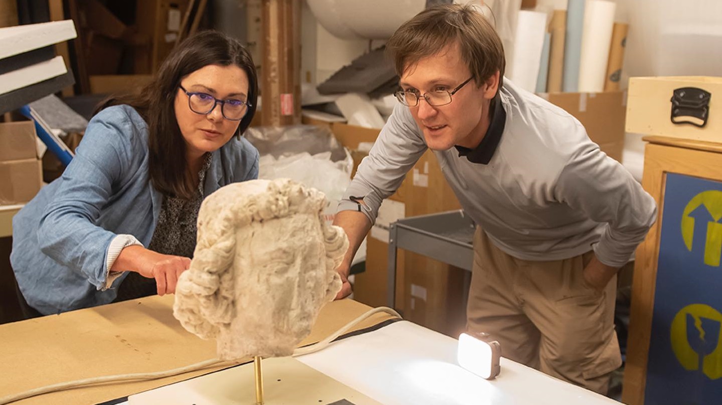 A woman and a man, both University of Alabama researchers, look at a sculpture of a head from Notre Dame in Paris inside a workroom.