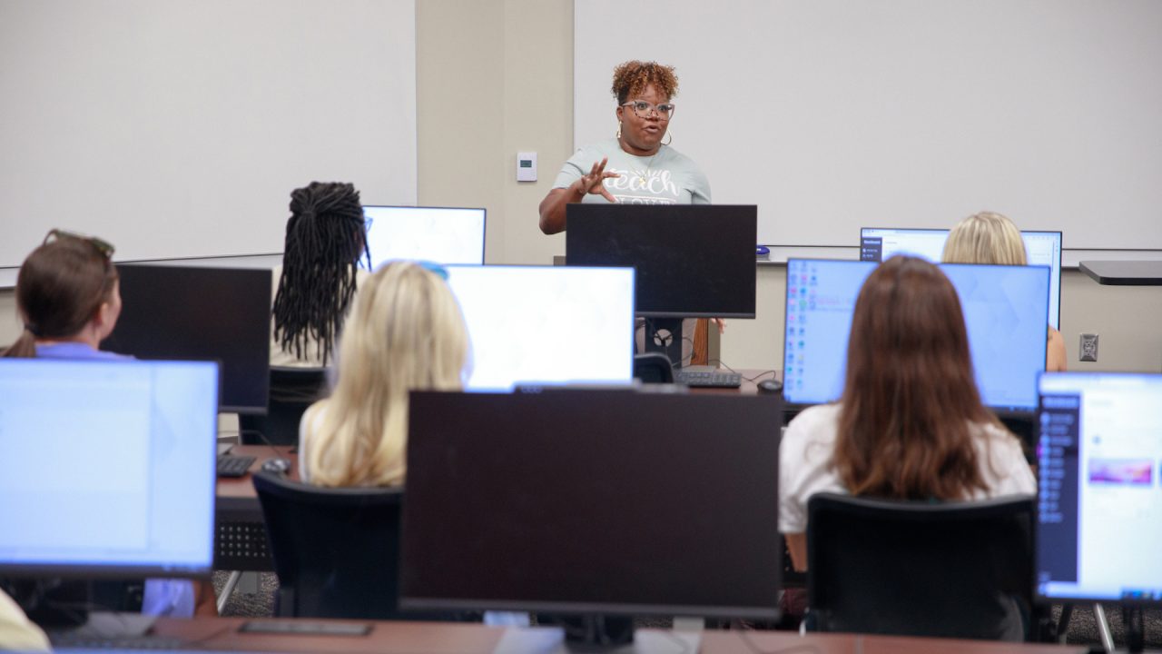 A woman teaches a course at The University of Alabama while students sit in front of computer screens.