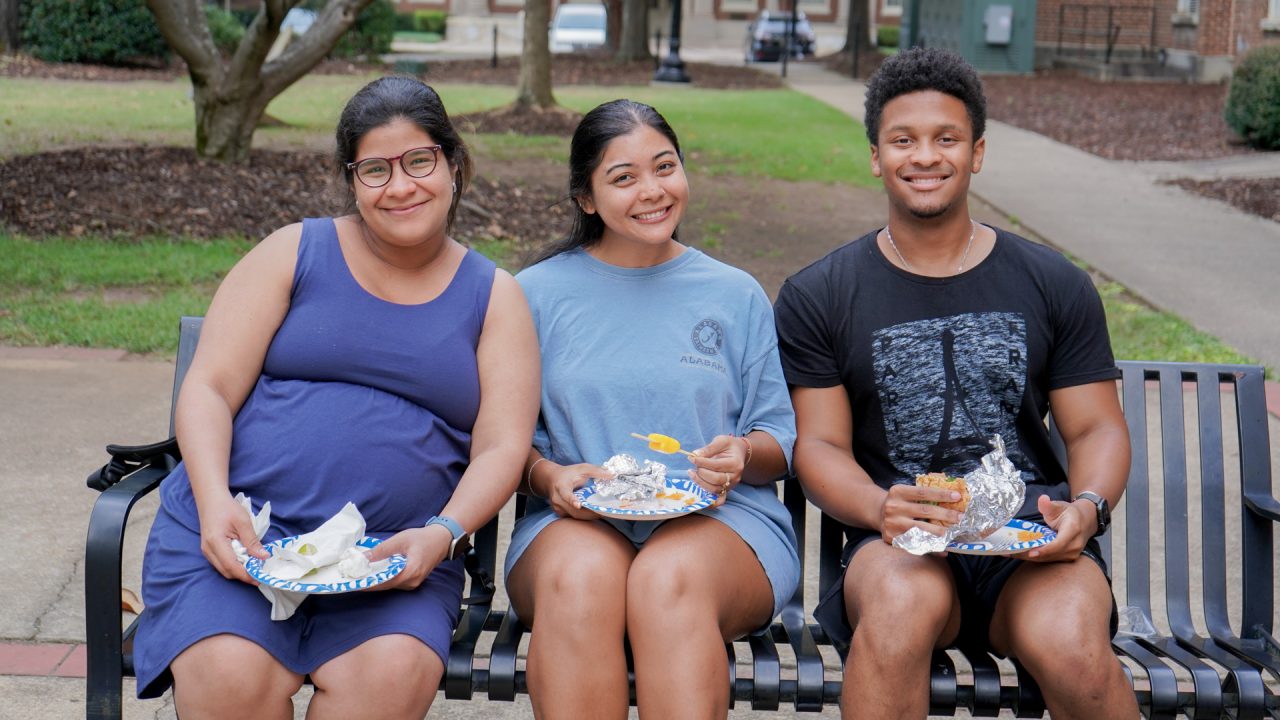 three students sit on a bench smiling while enjoying food