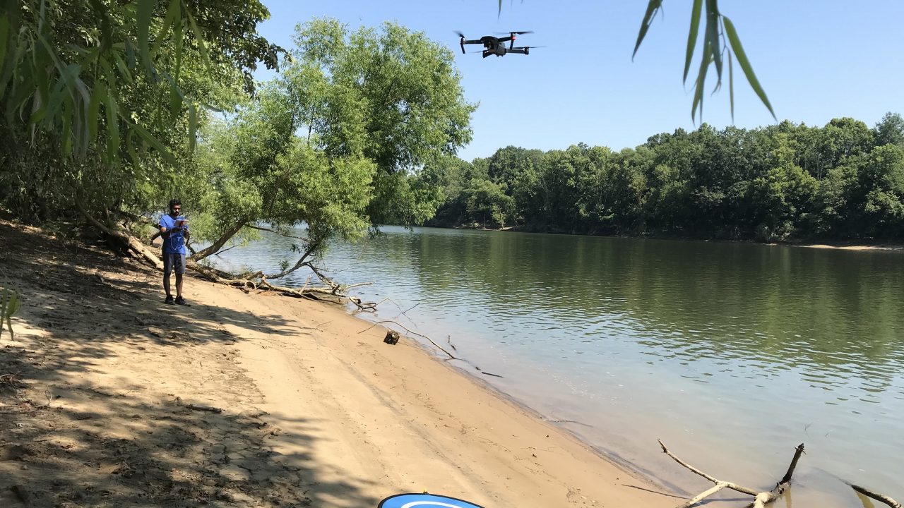 A man on the shore of a lake operate a drone flying overhead.