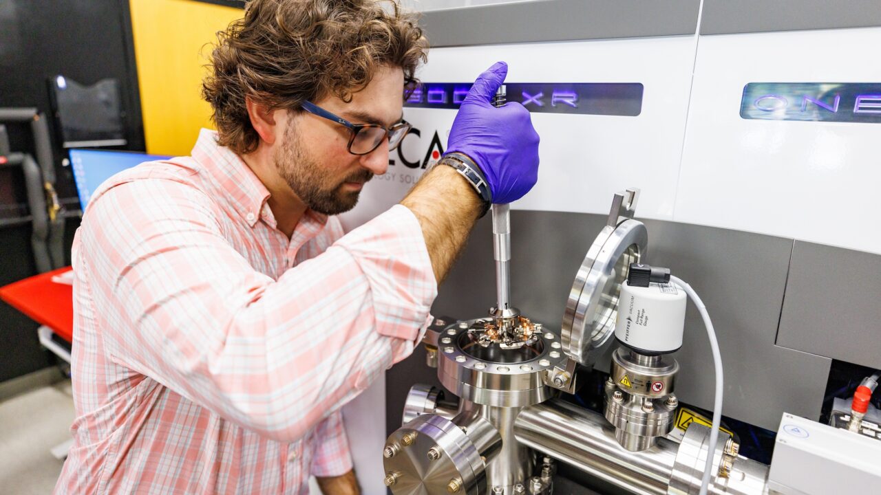 A man with gloves conducts materials research in front of metallic equipment.