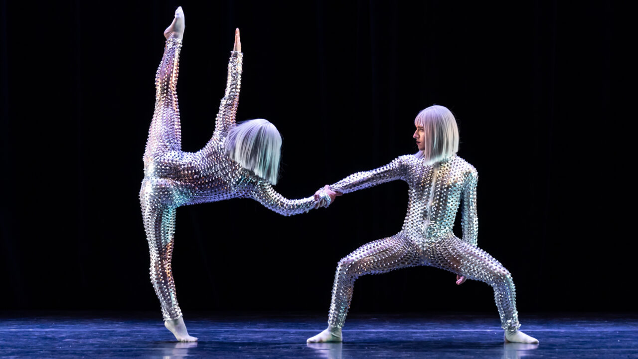 a dancer in a shiny metallic costume does a vertical split