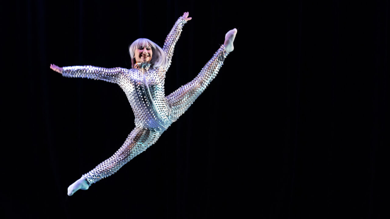 a female dancer in a shiny metallic costume leaps into the air