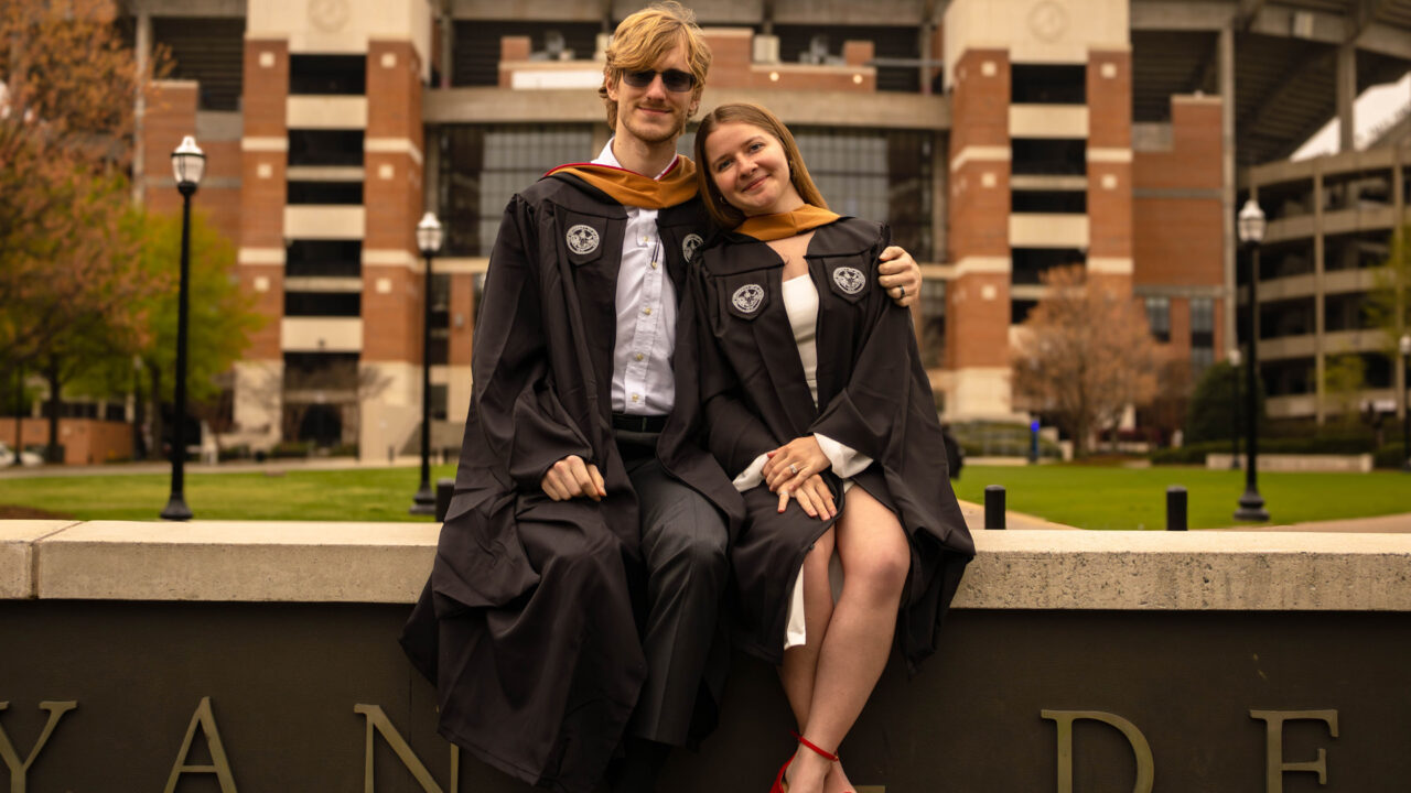 Jack and Hannah posing in their graduation regalia in front of Bryant Denny Stadium