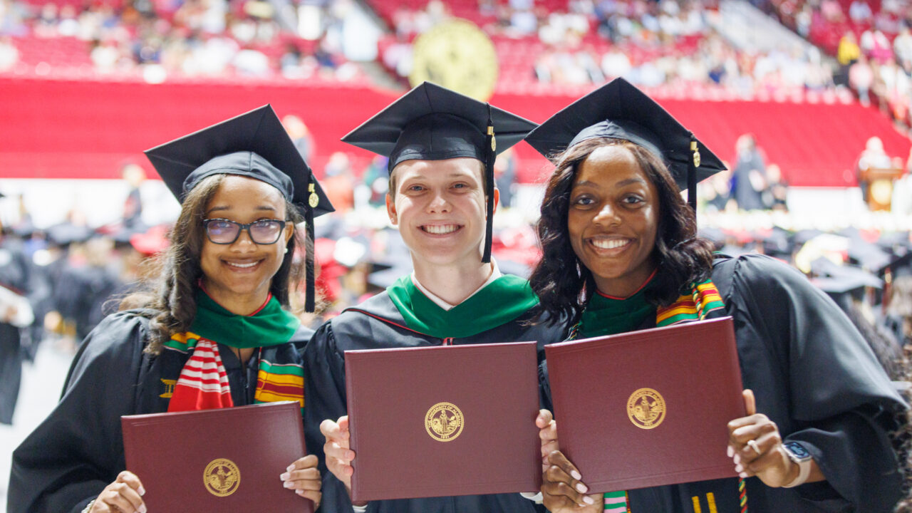 three new graduates smile into the camera holding up their degrees