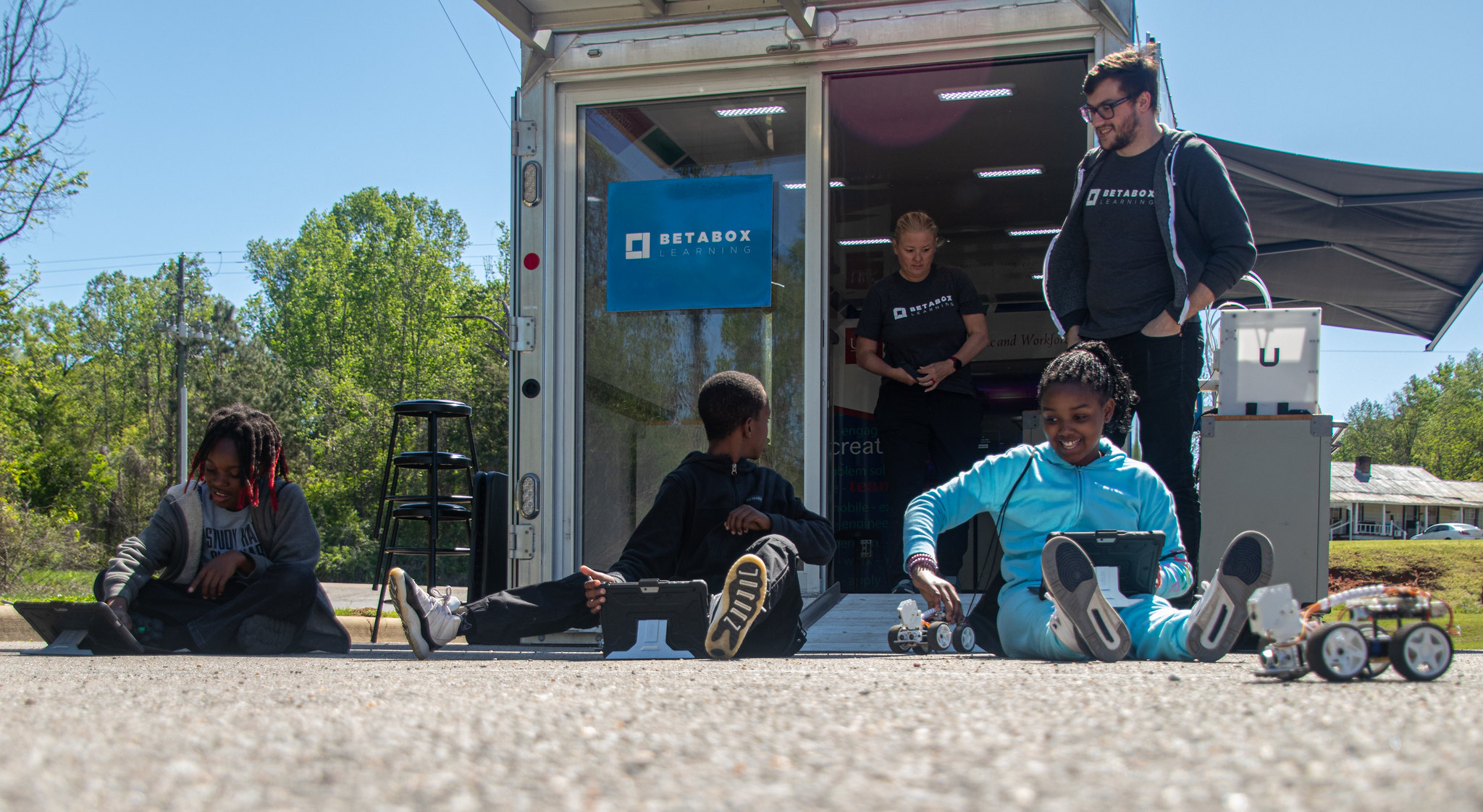 Three students sit on the ground as they operate 3-D printed remote control cars.