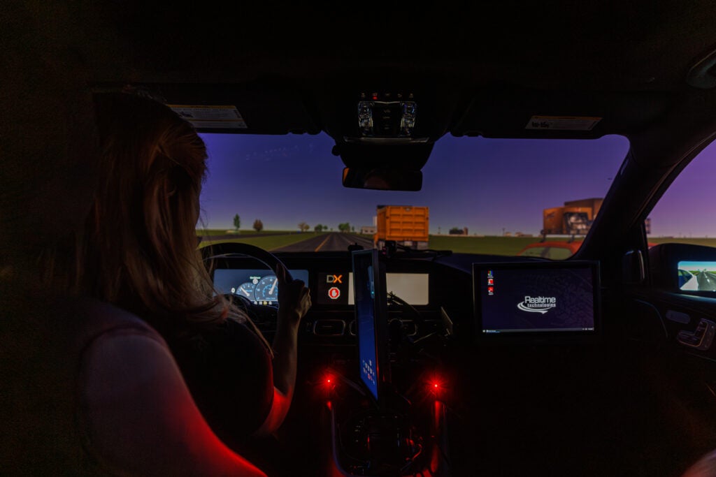 A driver sits inside a darkened vehicle with a large screen in front showing a road scene. Gauges are lit up inside the vehicle. 