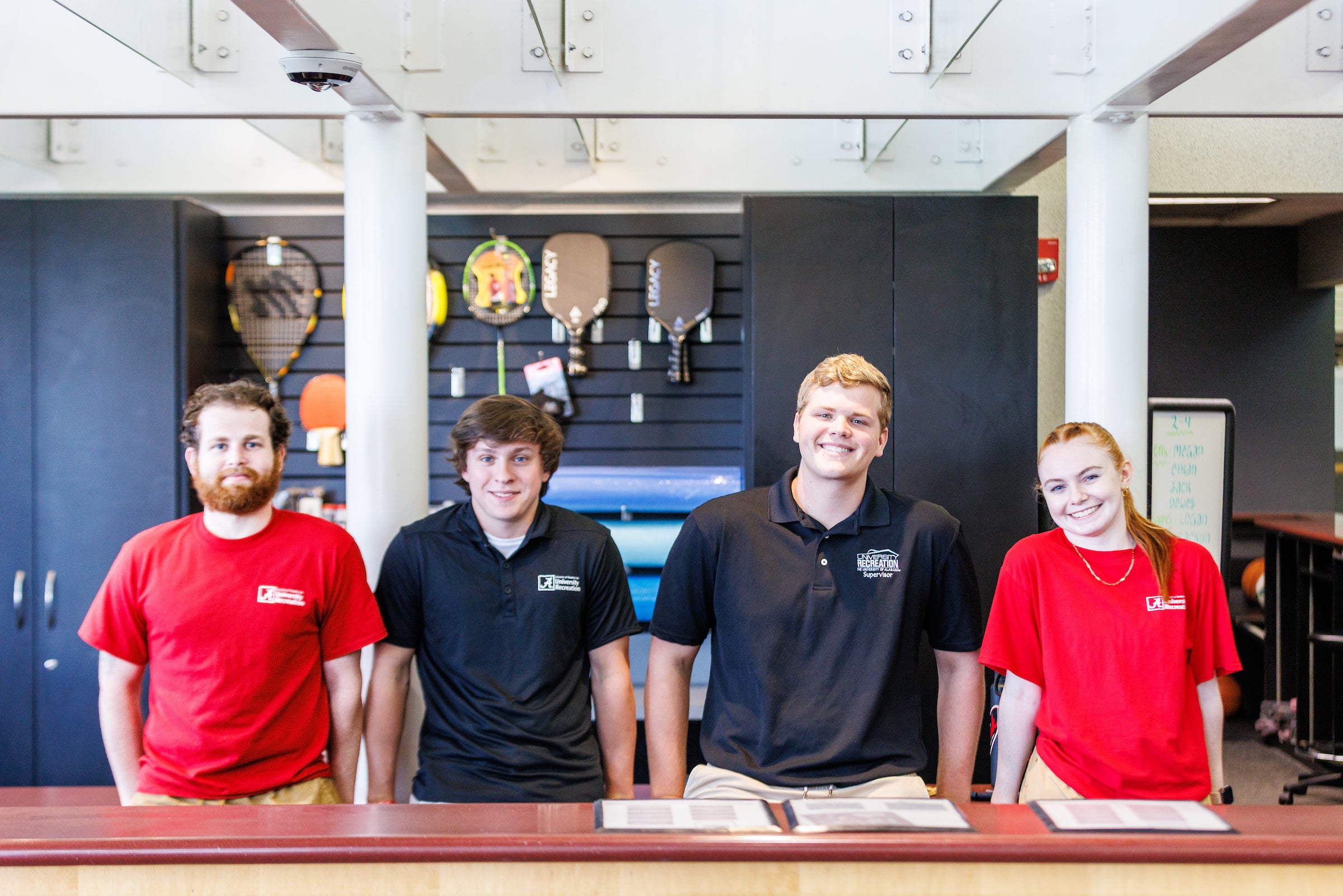 four students smile at the camera behind the front desk of the student recreation center