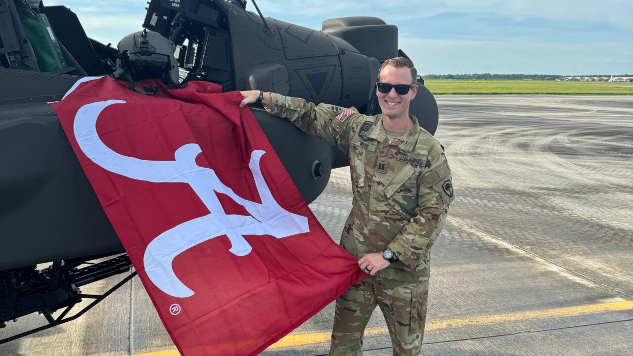 Kyle Wise stands next to an Apache helicopter while holding a flag with the Alabama Script A on it.