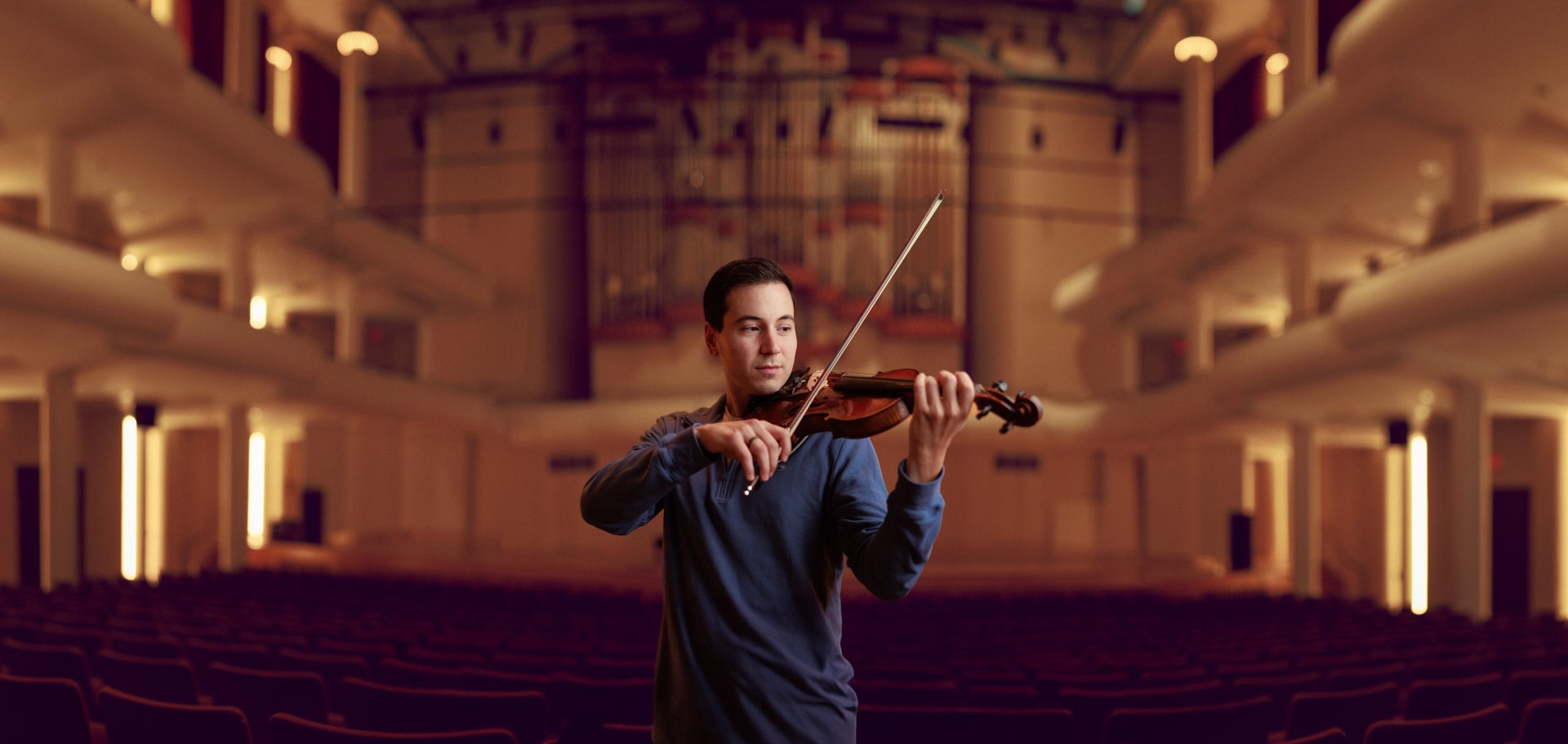 a male student plays a violin with the empty auditorium behind him