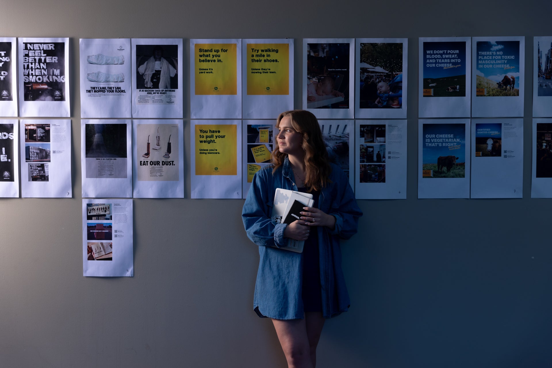 a student leans again a wall that is covered with print outs of graphic design work 