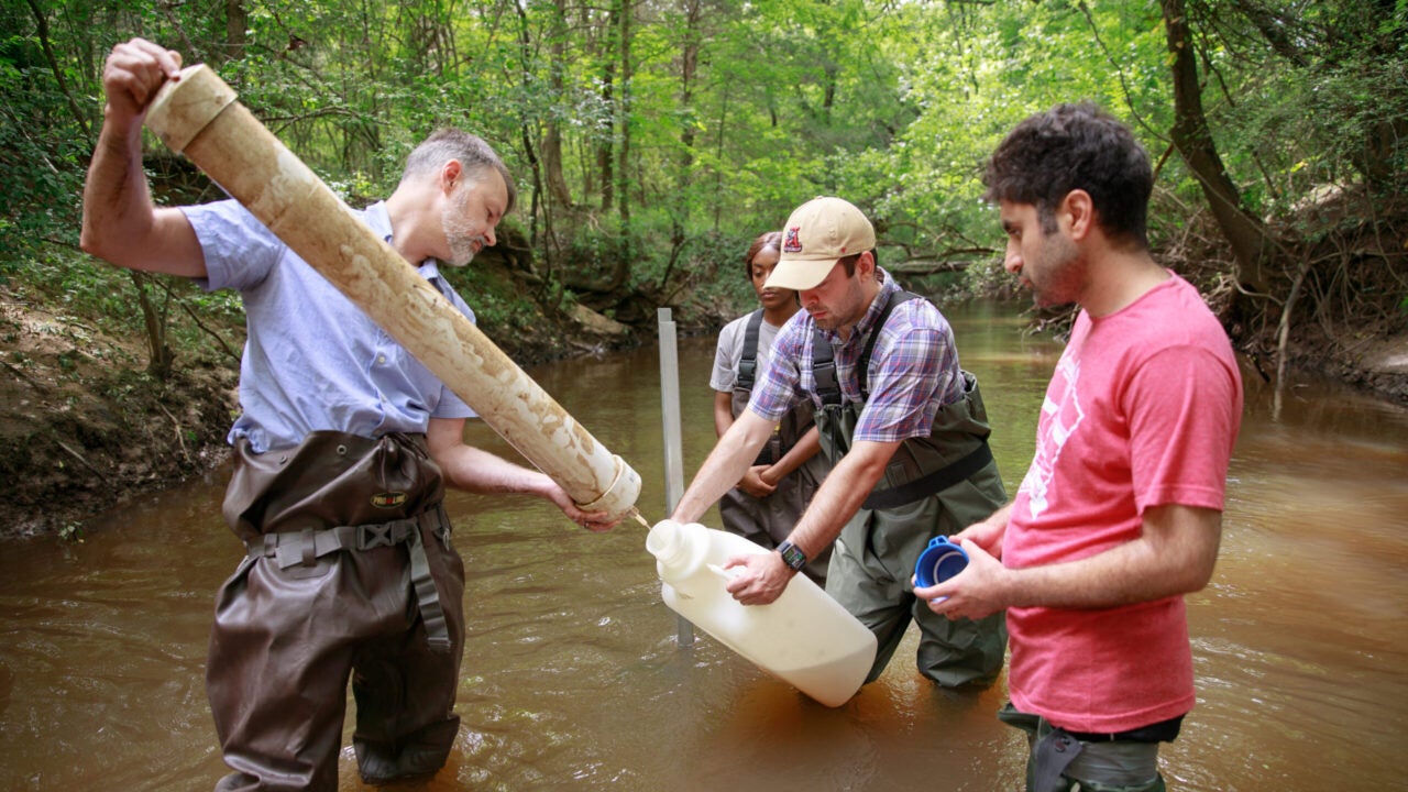 Dr. Mark Elliott and two students stand thigh-deep in an Alabama stream with instruments to collect water for water quality research.