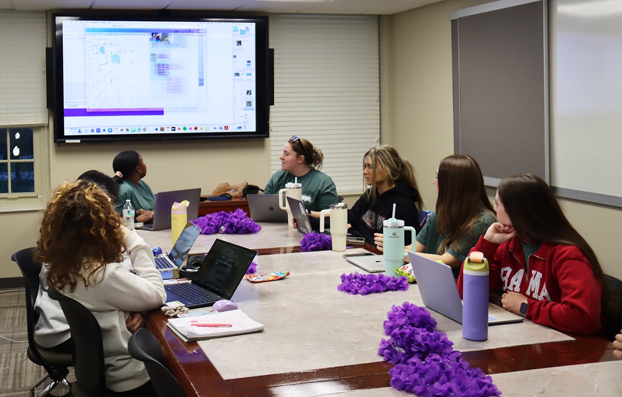 A small of group of students sit at a table and watch a presentation.