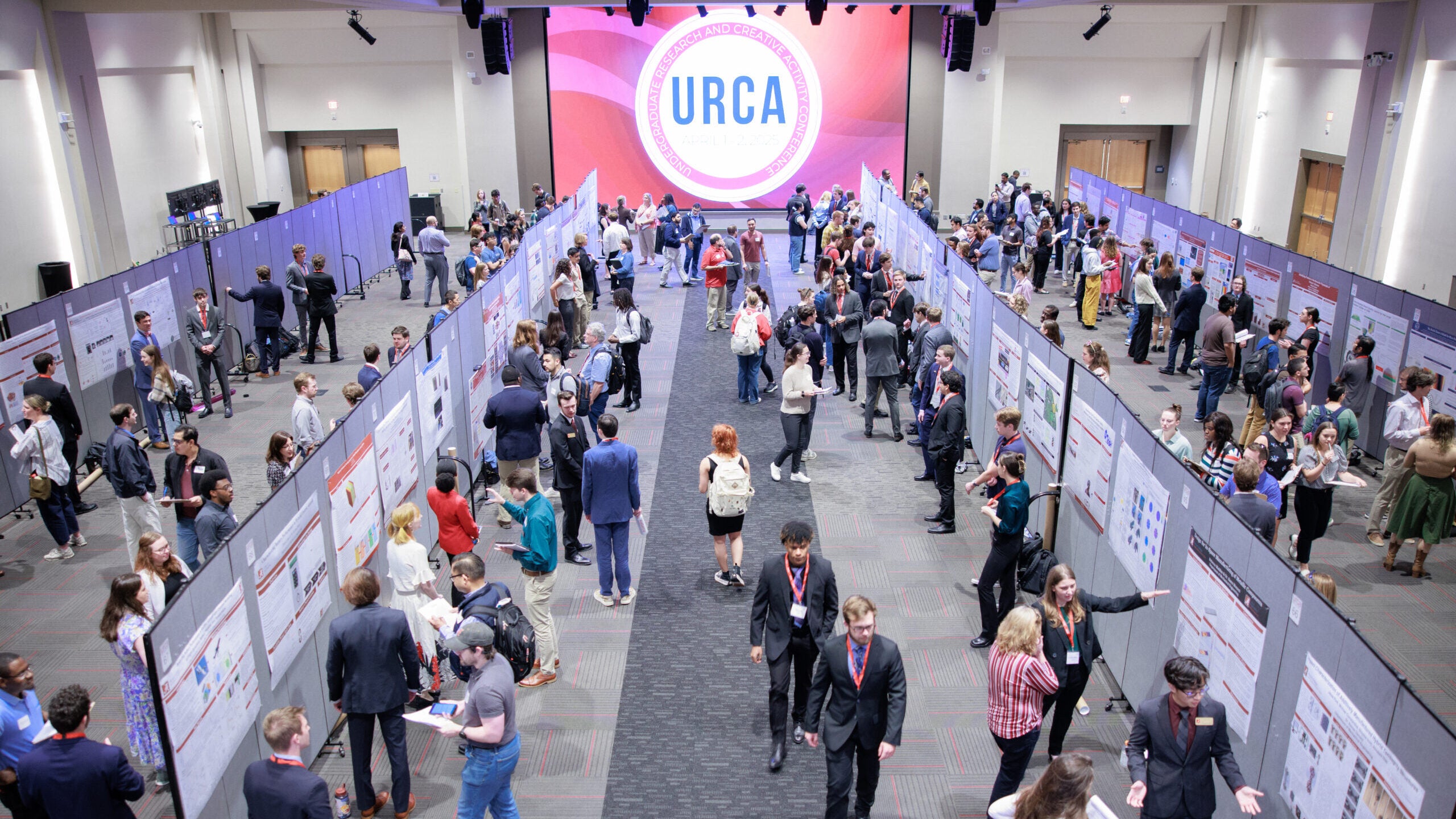 Students and faculty walk among several aisles with displays set up for research poster presentations in a large room. A banner at the end of the room reads URCA.