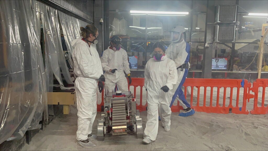 Members of the UA lunar robotics team wear white jumpsuits and masks in the competition space.