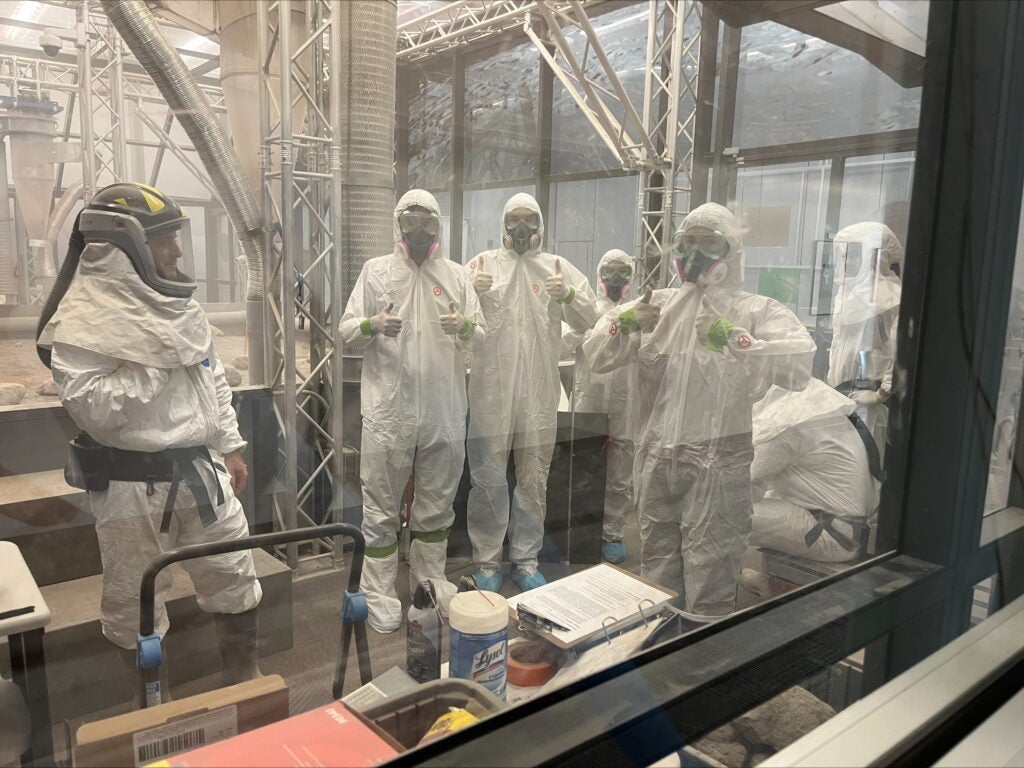 Four members of the UA lunar robotics team wear white jumpsuits and particle masks and show the thumbs-up signal.