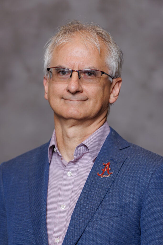 Dr. Konstantin Matchev in a blue blazer in front of a gray background. He is wearing a pin with the logo of the University of Alabama on his lapel.