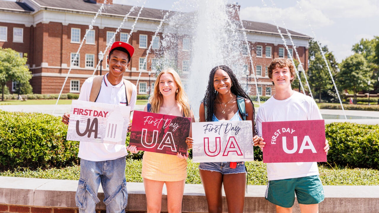 four students stand in front of the shelby fountain holding signs that say "first day UA"