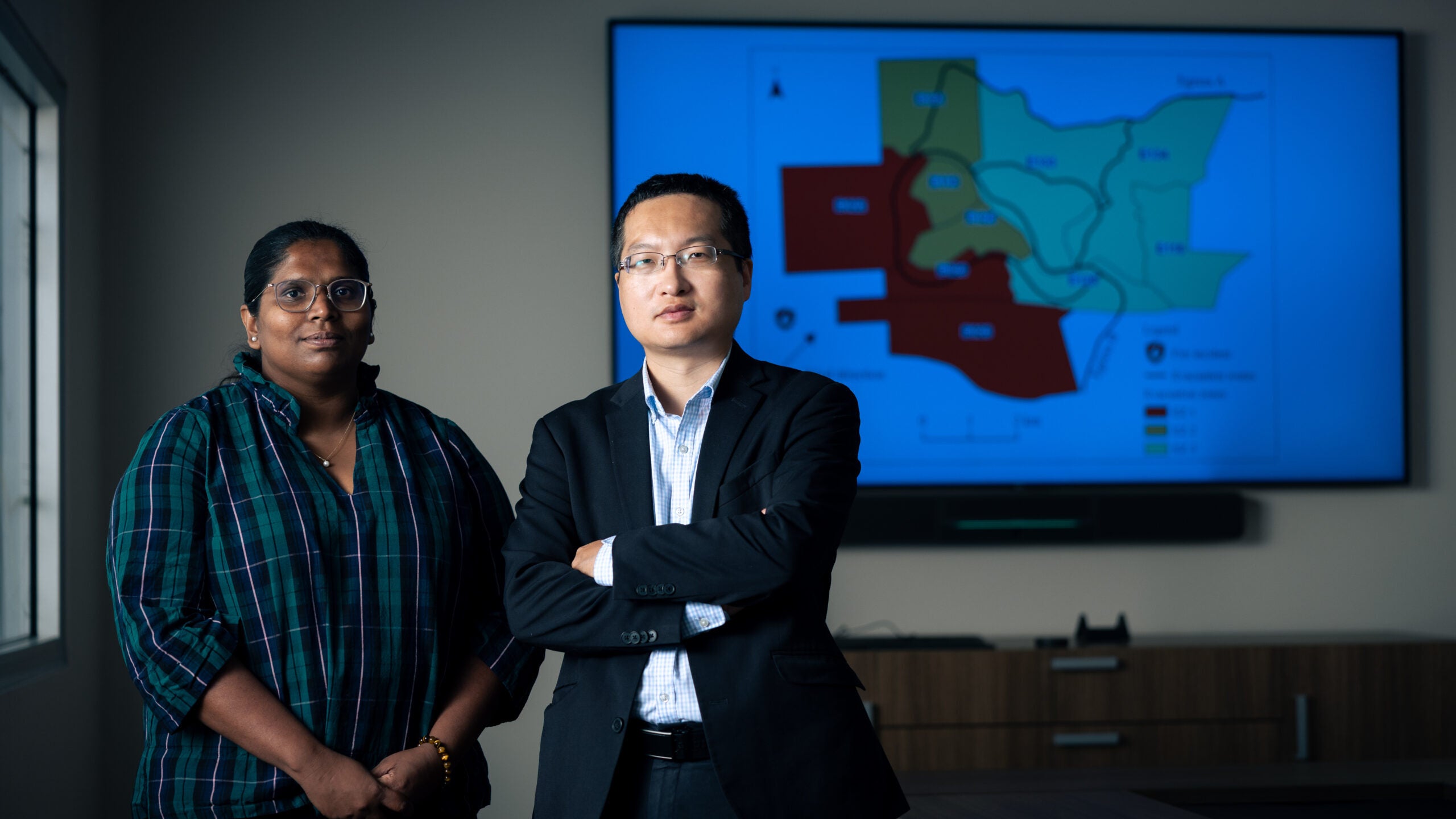 Two researchers, a man and a woman, stand in front of a large TV showing a map divided into brightly colored sections.