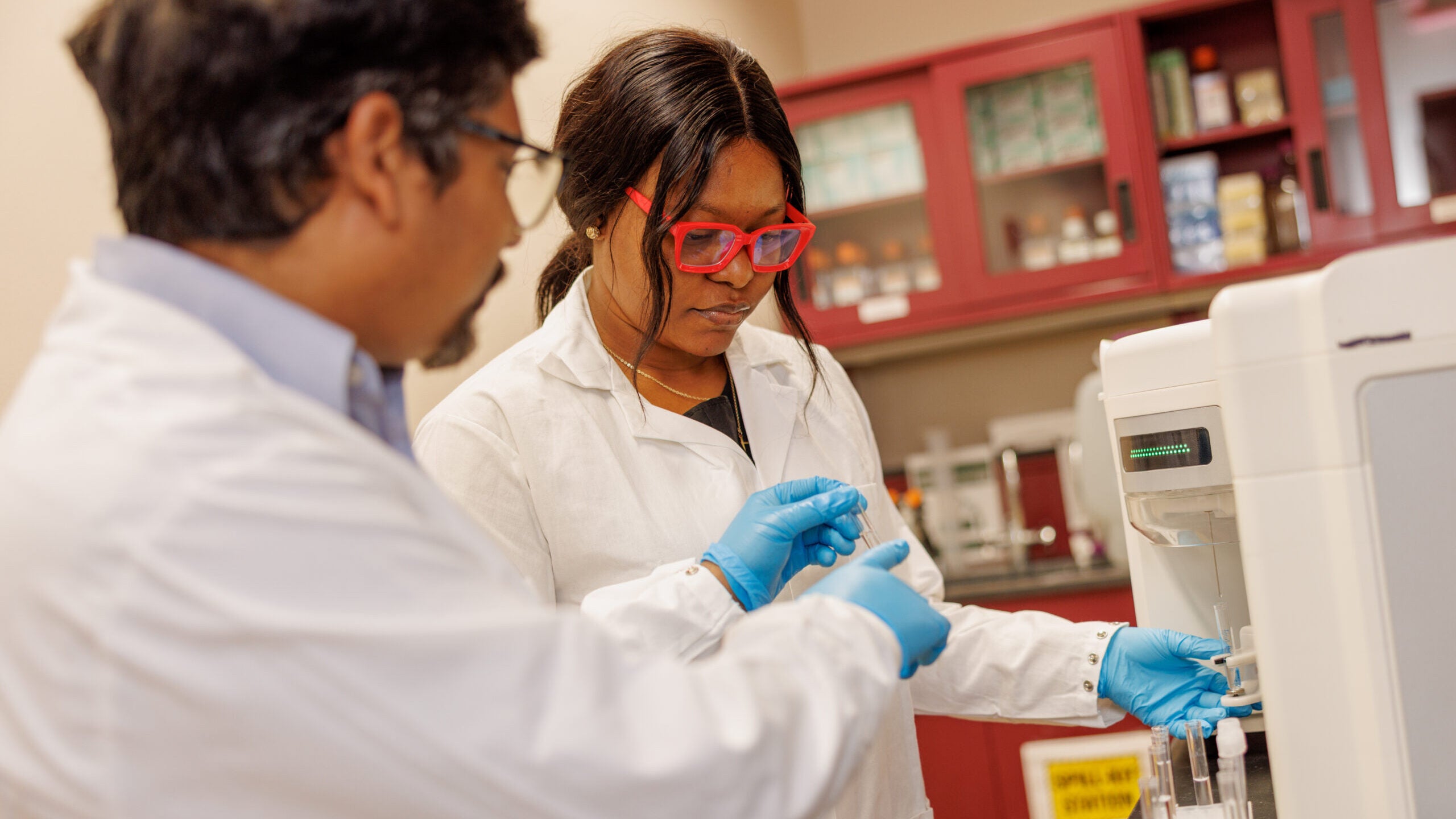 A man and a woman in white lab coats and blue vinyl gloves handle glass vials in a laboratory.