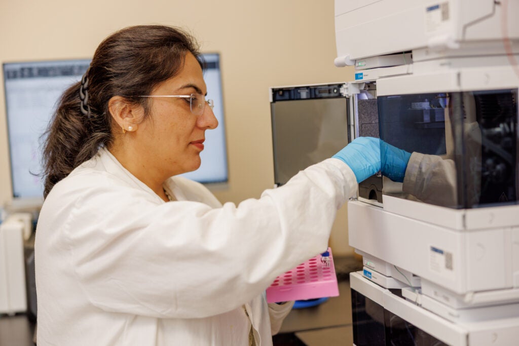 Dr. Meenakshi Arora, wearing a lab coat and blue vinyl gloves, putting samples into a machine in her lab at the University of Alabama