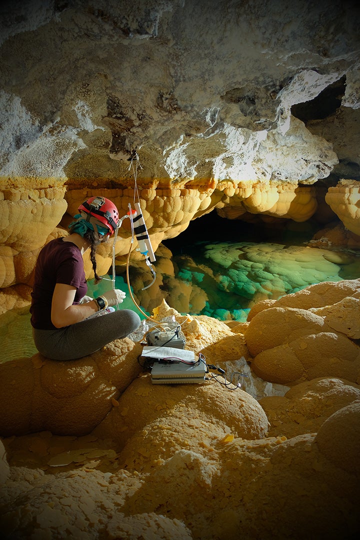 Graduate student Katey Bender collects biological samples from Lake Castrovalva in Lechuguilla Cave, New Mexico. A student wearing caving gear sits on yellow rocks beside a blue-green pool.