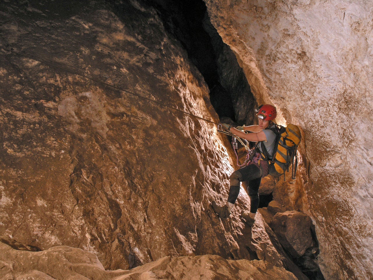 Hazel Barton, wearing a backpack and head lamp, crosses a sheer face of cave wall holding onto a rope with her boots braced against the rock.