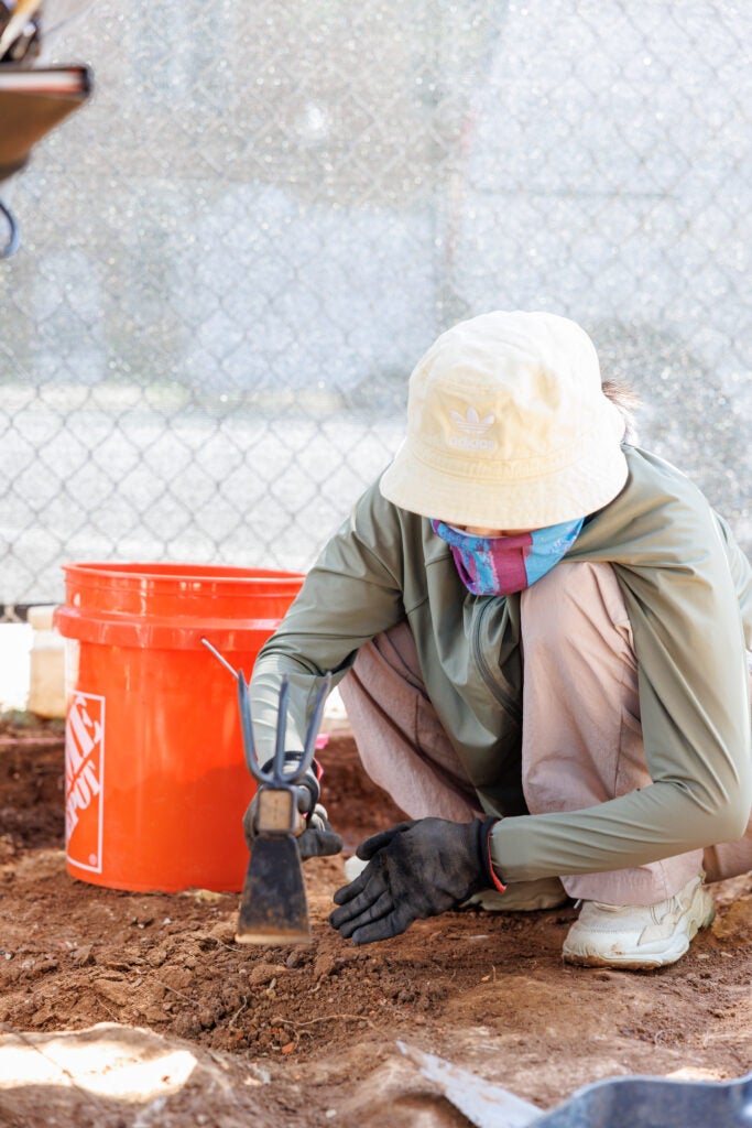A student at the archaeological field school wearing long sleeves and long pants, with a bucket hat and a scarf over their face, squats in the dirt beside an orange bucket. 