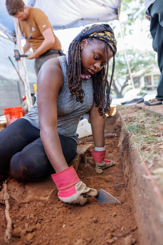 A female student wearing work gloves scrapes at a layer of dirt with a metal trowel at the archaeological field school site. 