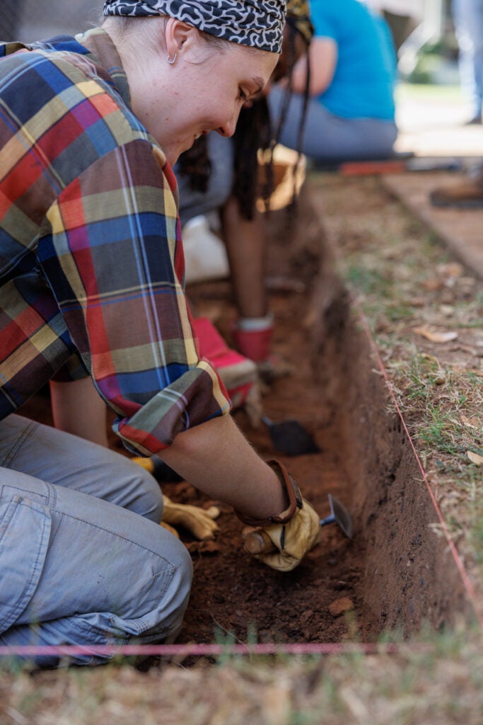 A student wearing a plaid shirt and yellow work gloves digs with a trowel at the archaeological field school. 