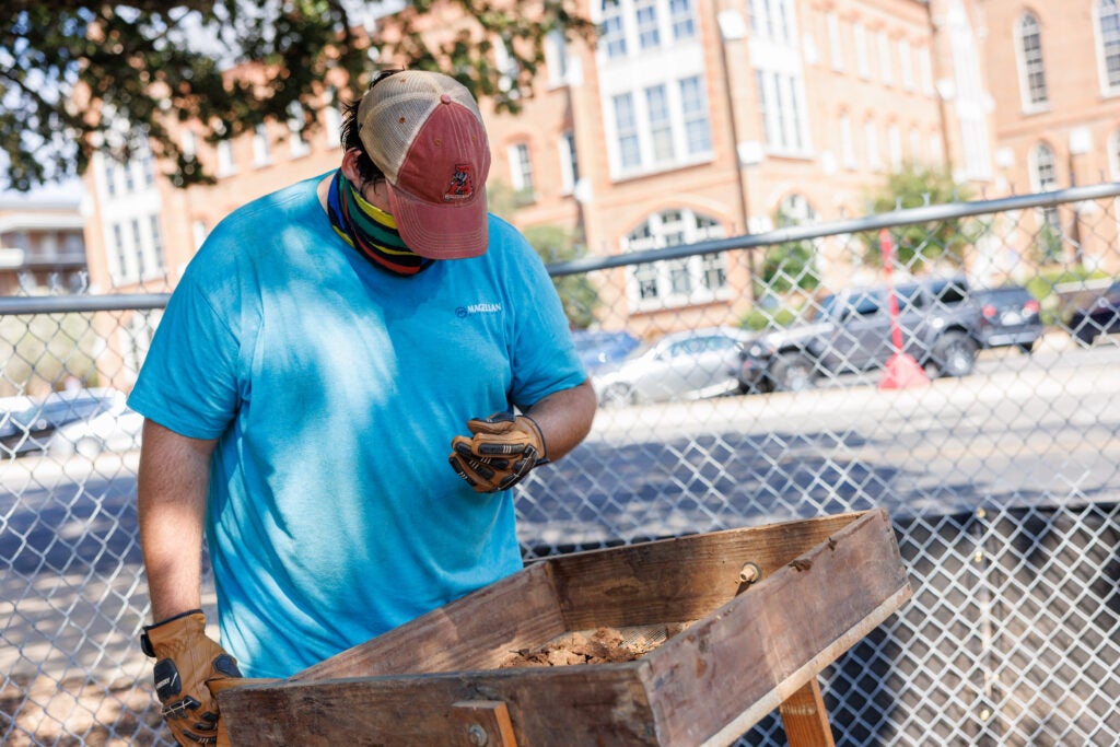 A male student at wearing a bandana over his mouth and nose stands at a wooden contraption with a screen to sift through dirt for smaller artifacts fragments. 