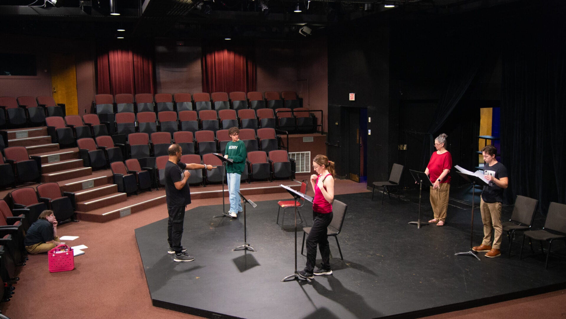 four students and a director rehearse a theatrical performance on a dark stage.