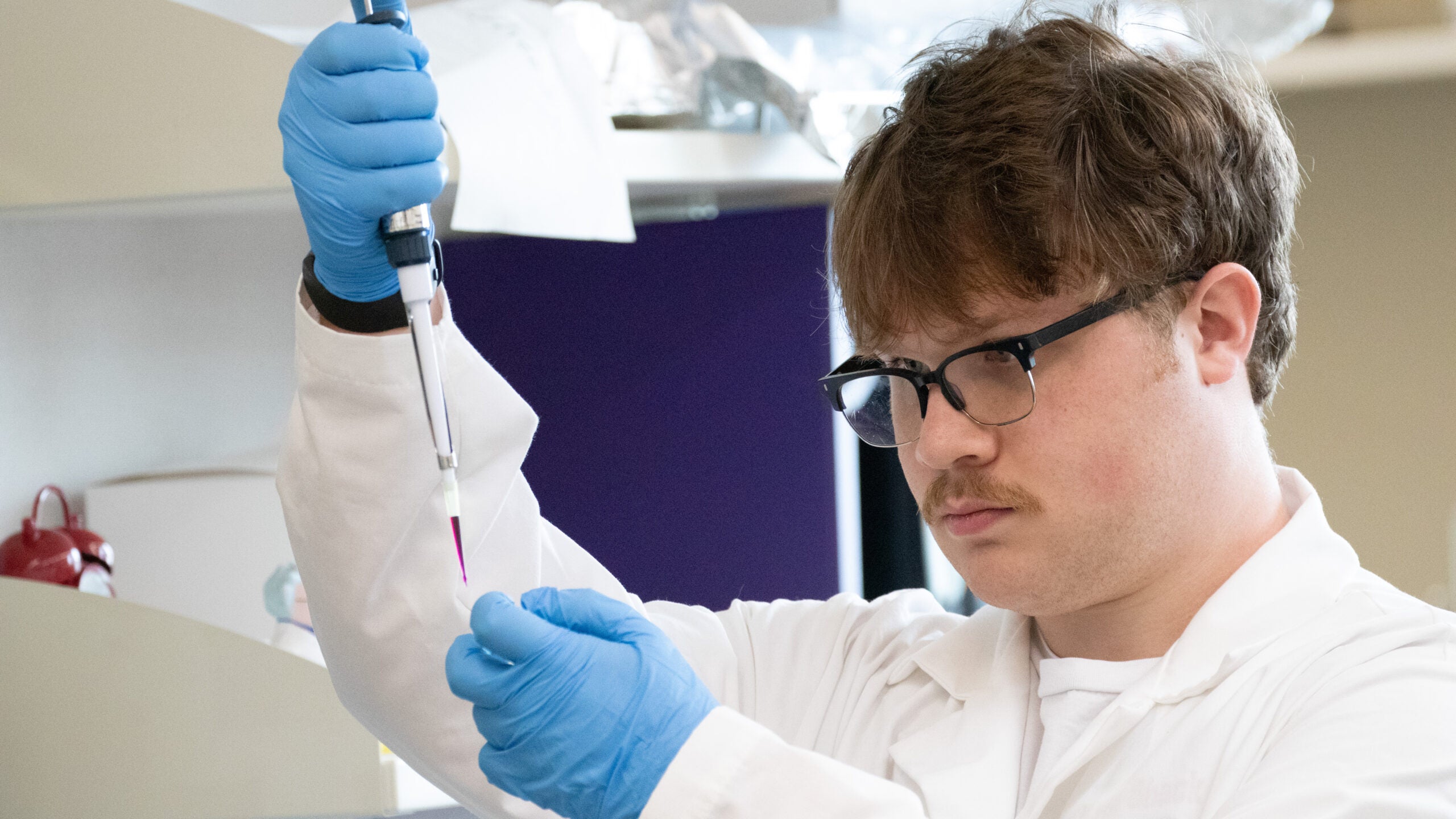 Aidan Meyers in the Caldwell Lab wearing a lab coat and holding a pipette with purple fluid.
