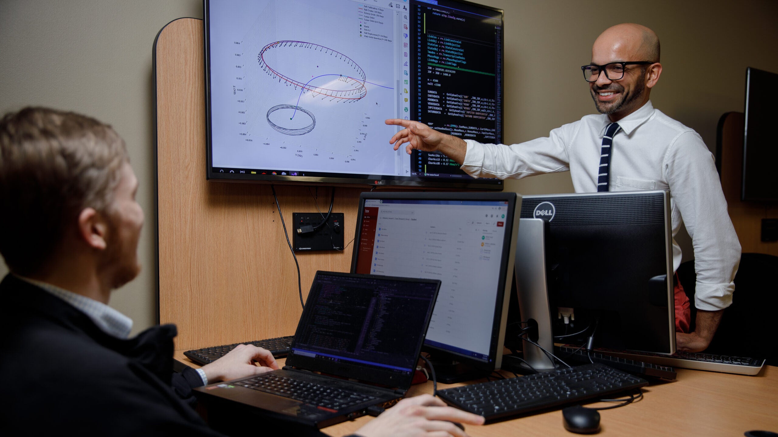 Dr. Rohan Sood points to a wall mounted monitor showing a diagram of a space orbit.