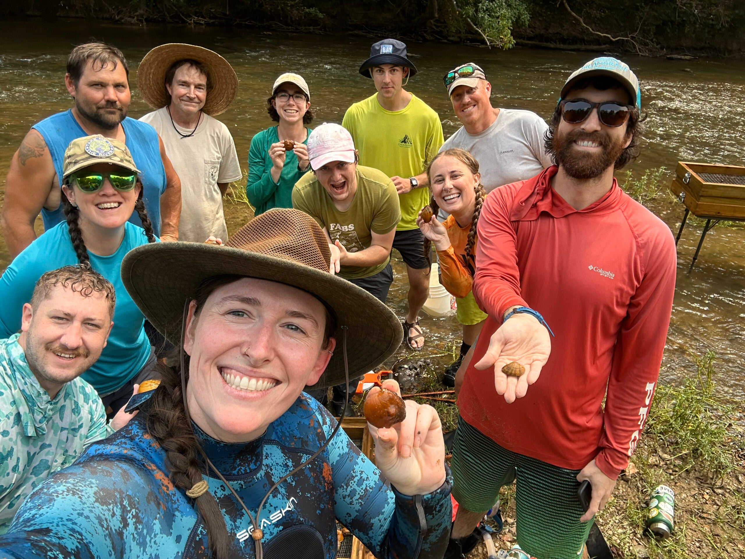 a group of men and women standing in front of a creek holding up freshwater mussels. 
